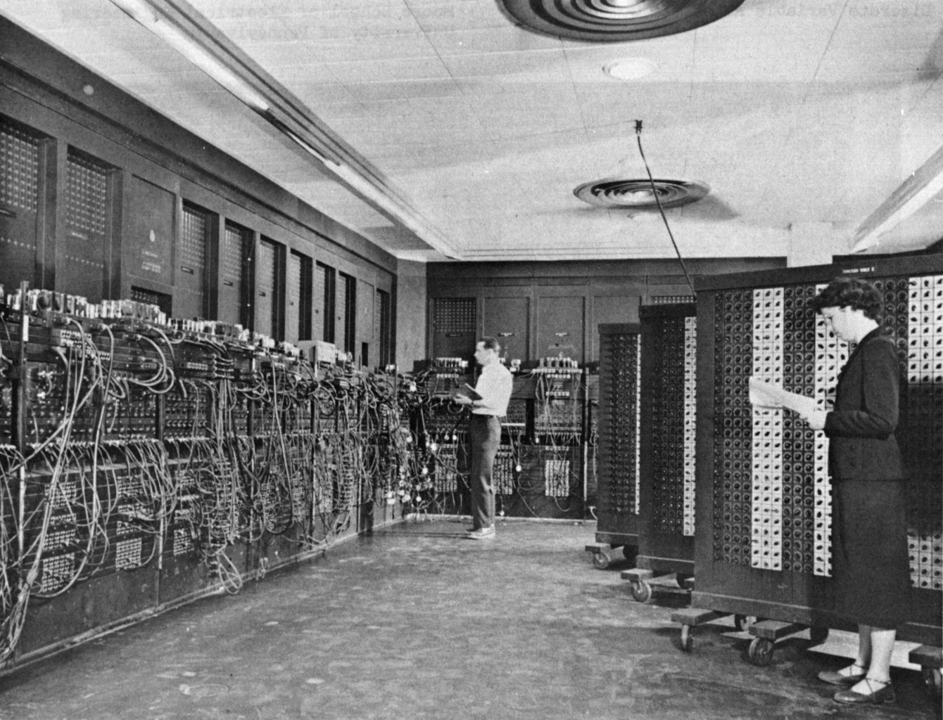 "A black and white photograph captures a large room filled with the tall electronic panels and tangled patch cables of the ENIAC computer system. Two people are positioned among the equipment, including a woman in the foreground standing beside a large control cabinet while holding a document."